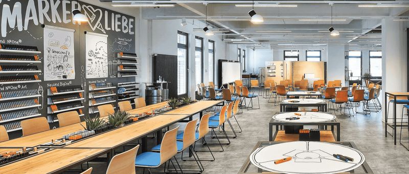 Geräumiger, moderner Workshop-Raum mit langen Tischen, Stühlen, Kunstzubehör und großen Fenstern. An den Wänden befinden sich kreative Designs und Regale mit Stiften.