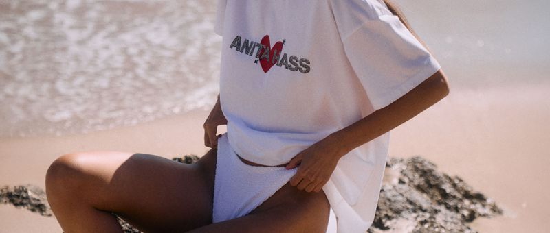Person, die am Strand sitzt und ein weißes T-Shirt mit der Aufschrift „ANTI” und einem roten Herzen sowie eine weiße Hose trägt. Wellen und Sand sind zu sehen.