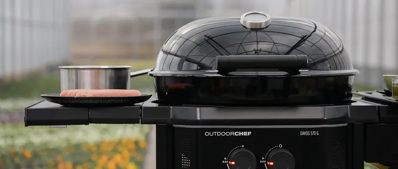 Ein schwarzer Outdoor-Grill mit gewölbtem Deckel, ein Hotdog auf einem Teller und ein Topf auf der Ablage, in einem Gewächshaus.