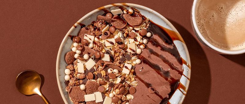 Eine Schüssel Schokoladengranola mit verschiedenen Toppings und eine Tasse Kaffee auf einer braunen Oberfläche, dazu ein goldener Löffel.