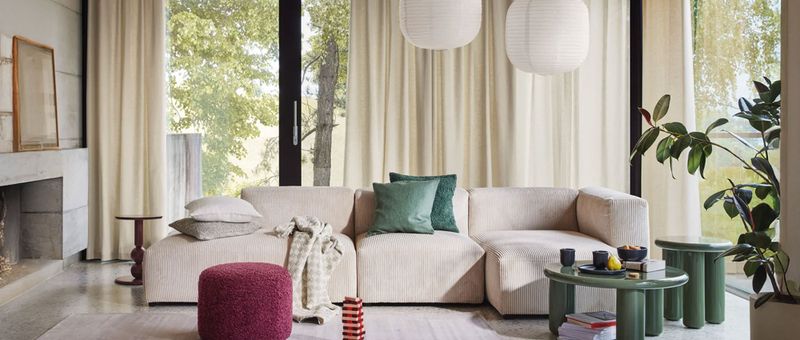 Modernes Wohnzimmer mit einem beigen Ecksofa, grünen und weißen Kissen, einer rosa Ottomane und einem grünen Couchtisch. Große Fenster und Pflanzen sorgen für viel Licht.