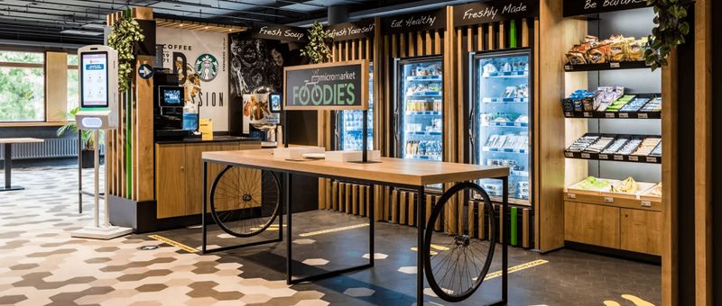Modernes Convenience-Store-Interieur mit Holzakzenten, Kühlregalen, Snack-Displays, einem Selbstbedienungskiosk und einem „Foodies“-Schild auf einem Tisch.