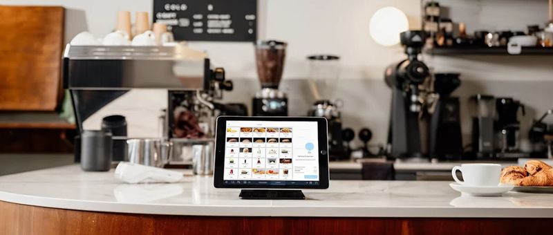 Auf einer Café-Theke steht ein Tablet mit einem Menü, im Hintergrund sind Kaffeemaschinen und eine Tasse mit Croissants zu sehen.