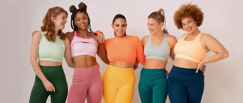 Fünf Frauen in farbenfroher Activewear lächeln und stehen zusammen vor einem neutralen Hintergrund und zeigen verschiedene Körpertypen und Stile.