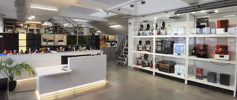 Ein modernes Ladeninnere mit 3D-Druckern in Regalen, einem schlichten weißen Tresen und einer Pflanze. Eine Treppe führt zu einem Zwischengeschoss mit weiteren Artikeln.