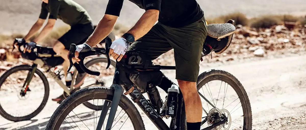 Zwei Radfahrer fahren mit Schotterfahrrädern auf einem steinigen Feldweg. Sie tragen legere Fahrradkleidung und Handschuhe, im Hintergrund ist eine verschwommene Wüstenlandschaft zu sehen.