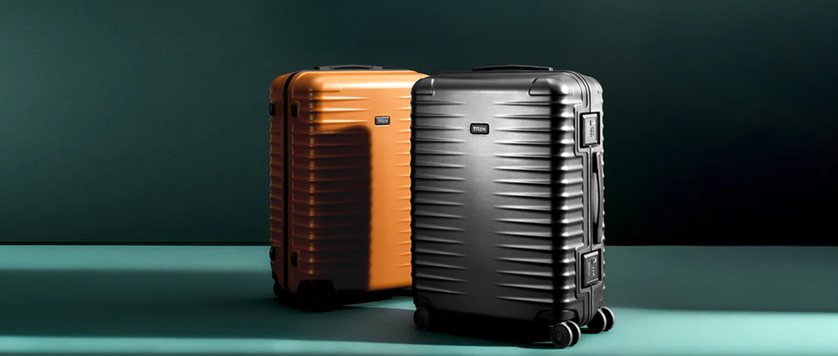 Zwei Hartschalenkoffer, einer orange und einer schwarz, stehen aufrecht auf einer grünen Fläche vor einem dunkelgrünen Hintergrund.