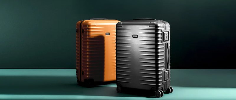 Zwei Hartschalenkoffer, einer orange und einer schwarz, stehen aufrecht auf einer grünen Fläche vor einem dunkelgrünen Hintergrund.