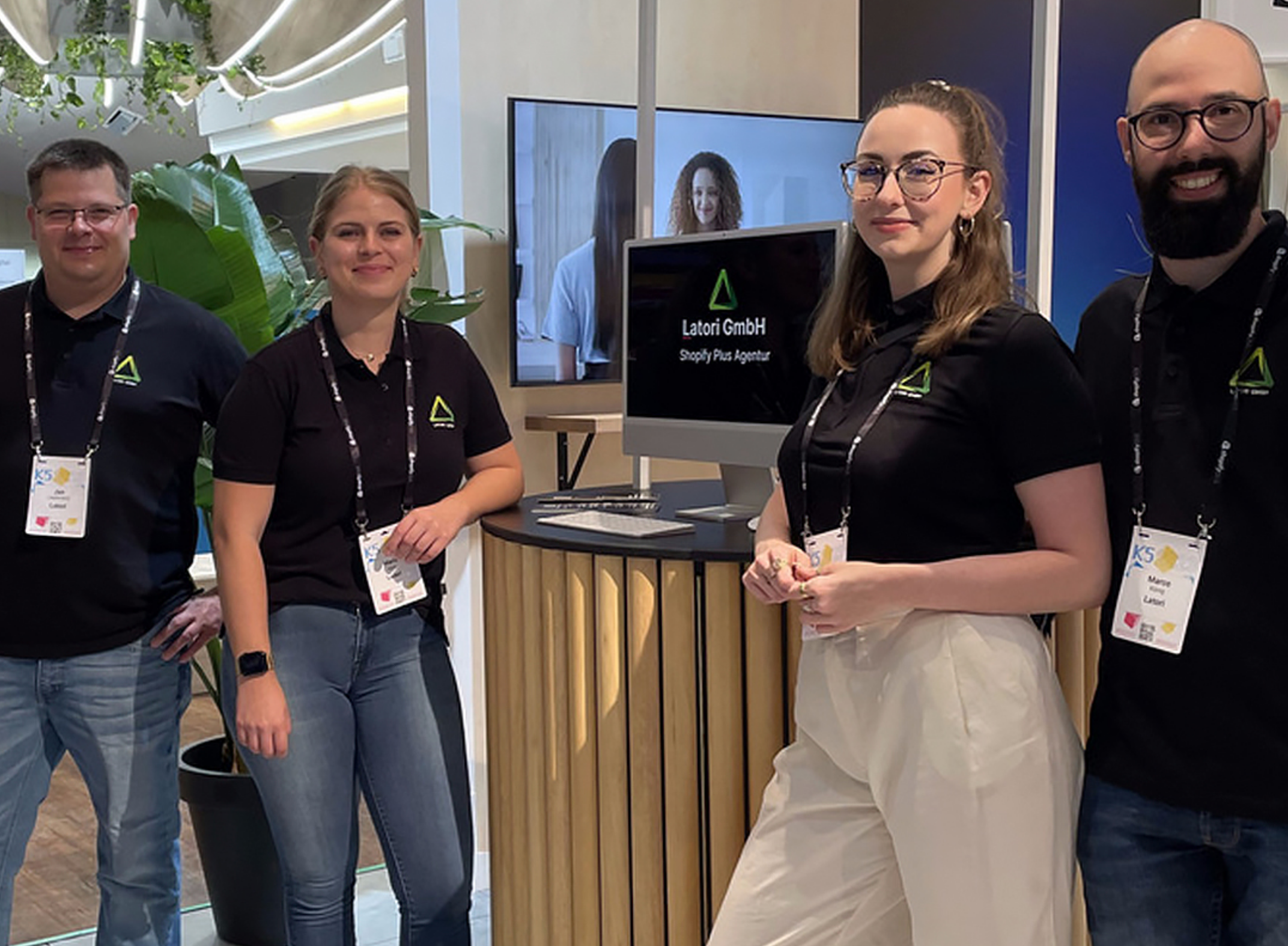 Vier Personen in Marken-Shirts stehen lächelnd an einem Messestand mit einem Monitor, auf dem „Latori GmbH“ zu sehen ist.