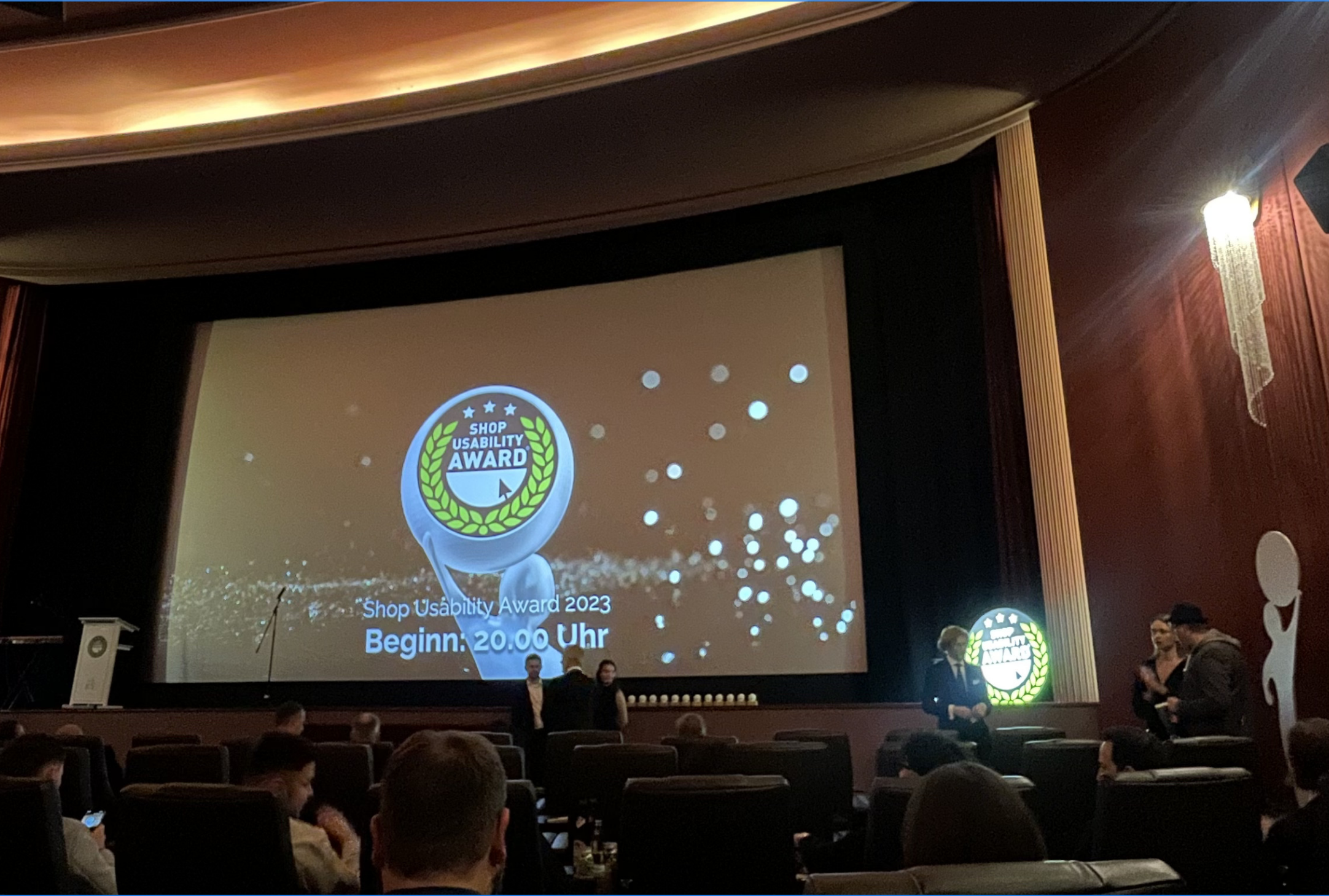 Ein Theater mit einer großen Leinwand, auf der „Shop Usability Award 2023” zu sehen ist. Die Zuschauer sitzen, einige stehen in der Nähe der Bühne.