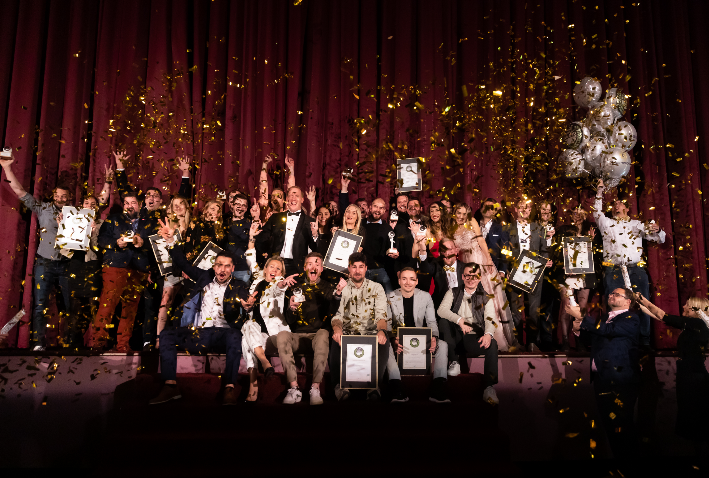 Eine Gruppe von Menschen feiert auf der Bühne mit Konfetti und Auszeichnungen, einige halten Luftballons in den Händen, vor einem roten Vorhang.