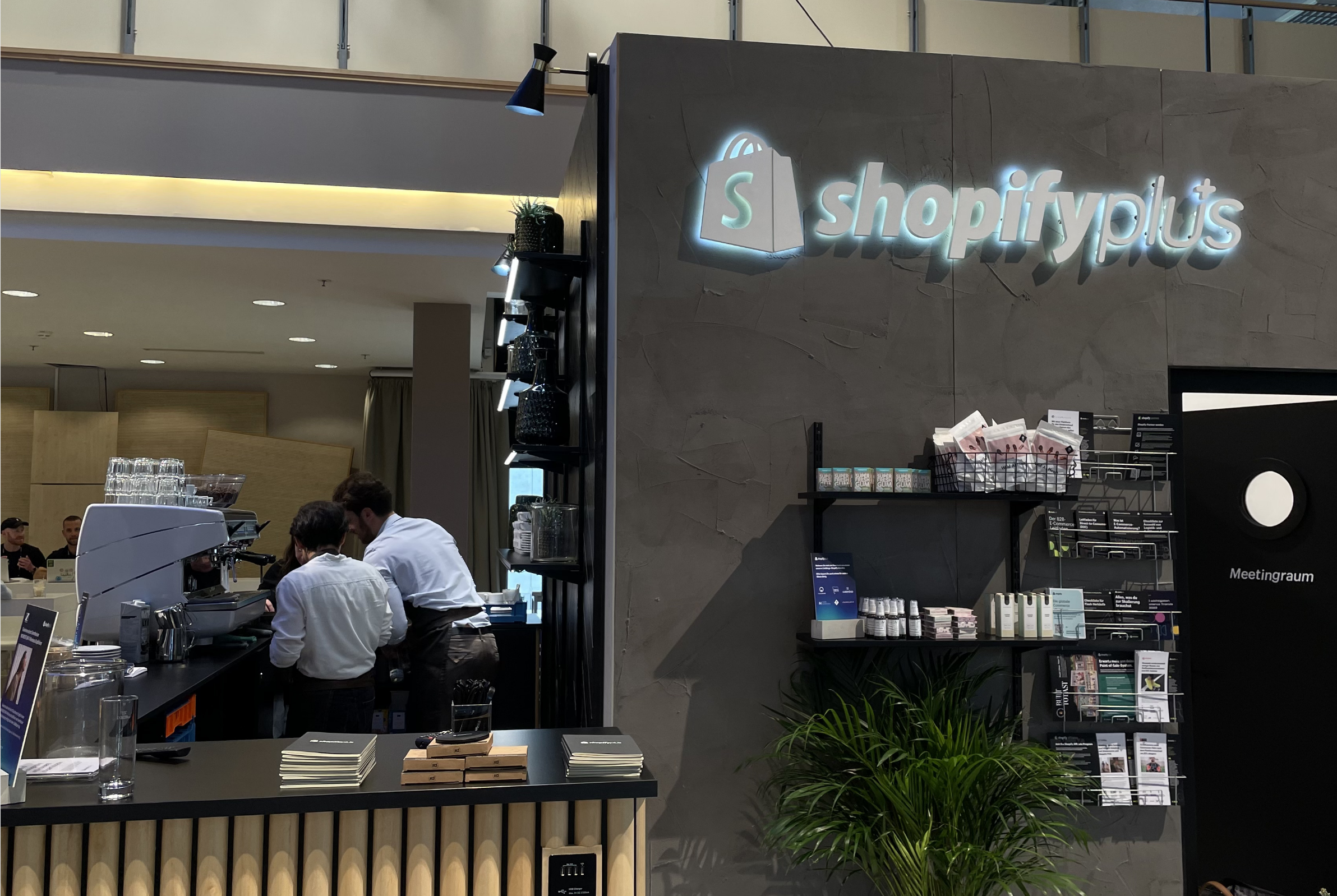 Innenraum eines Cafés mit zwei Baristas an der Theke. „Shopify Plus”-Schild an der Wand, Regale mit Produkten und eine Topfpflanze in der Nähe.