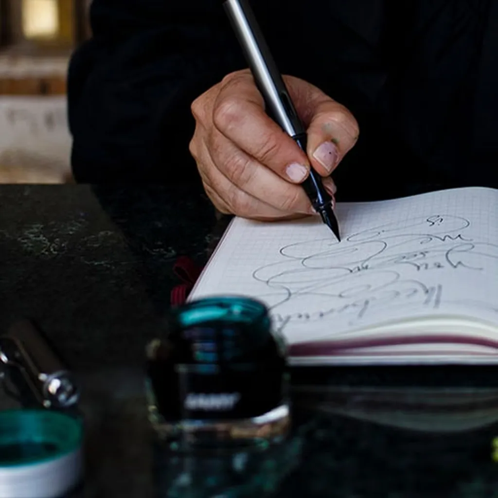 Eine Person, die mit einem Stift in ein Notizbuch schreibt, umgeben von Grünzeug und einem Tintenfass auf einer dunklen Fläche.