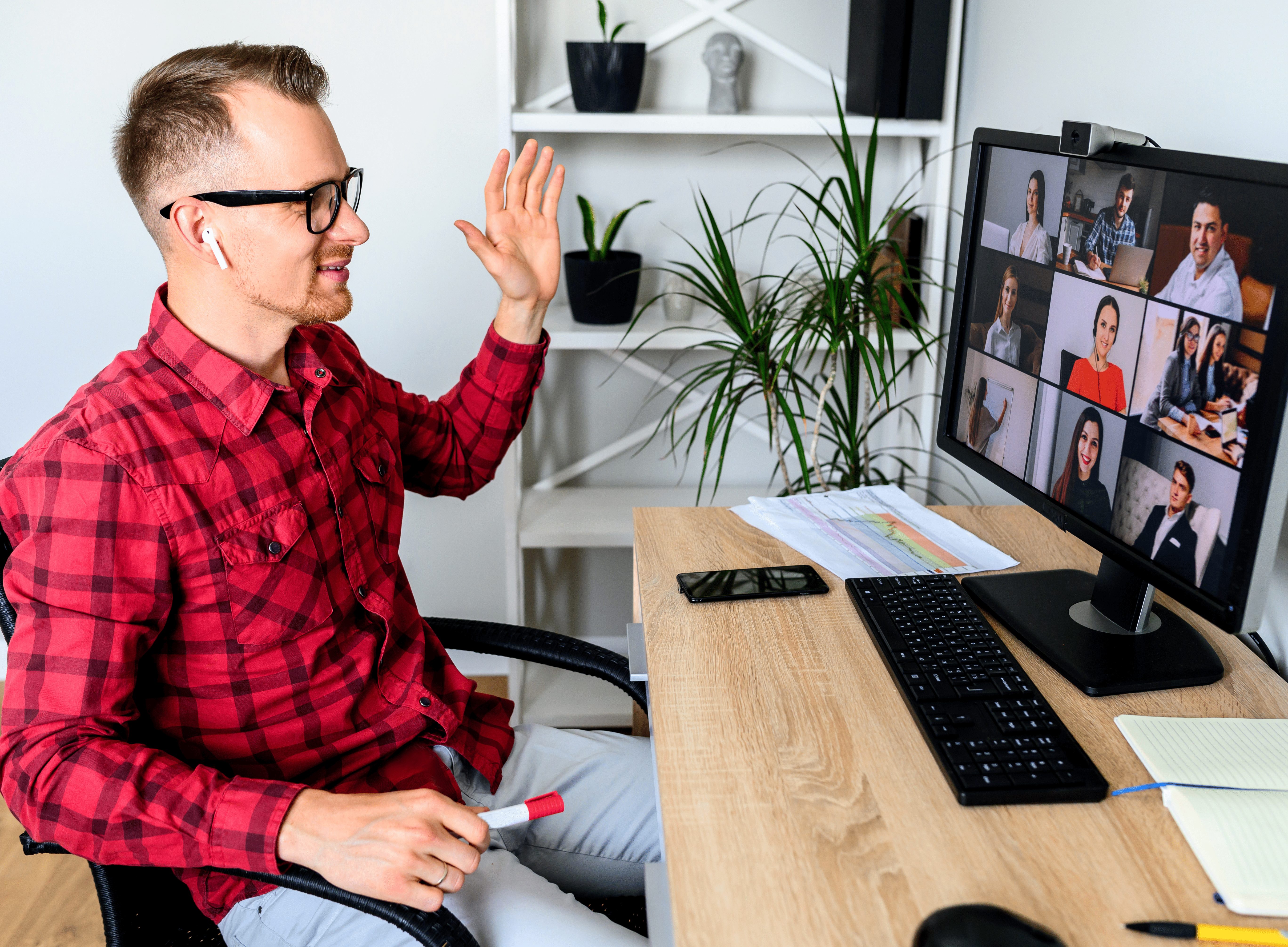 Mann im rot-karierten Hemd sitzt am Schreibtisch und winkt während einer Videokonferenz mit mehreren Teammitgliedern am Computerbildschirm.