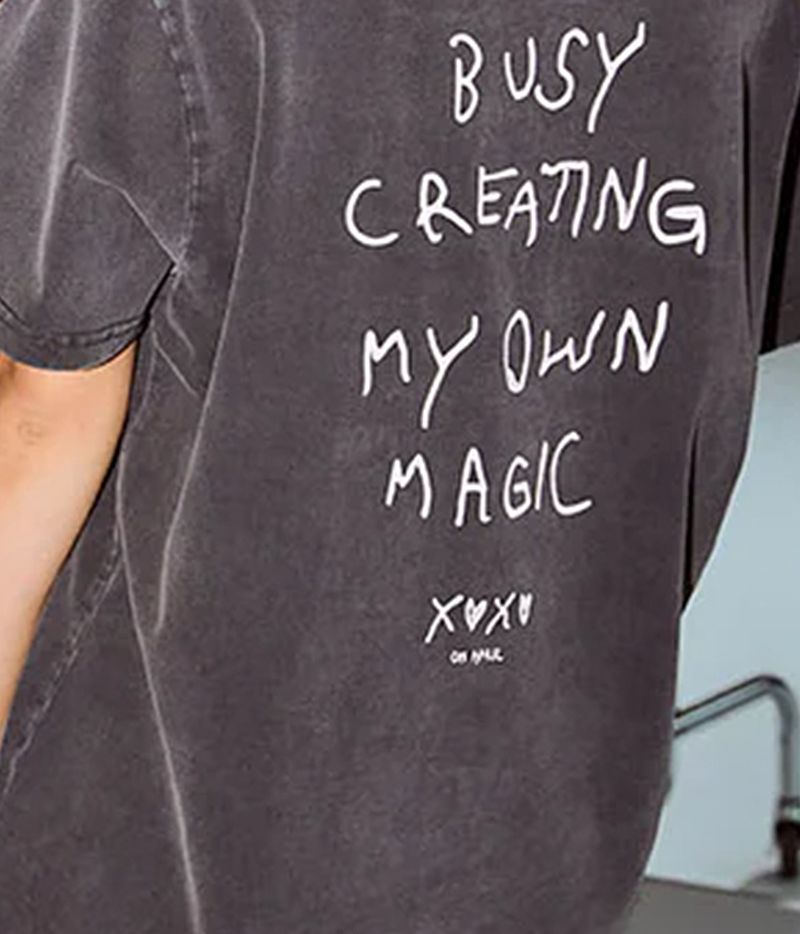 Person mit einem verblassten schwarzen T-Shirt mit der Aufschrift „Busy creating my own magic” (Beschäftigt damit, meine eigene Magie zu erschaffen) auf dem Rücken, stehend neben einem Kleiderständer mit Mänteln und einer Person in Burgunderrot.