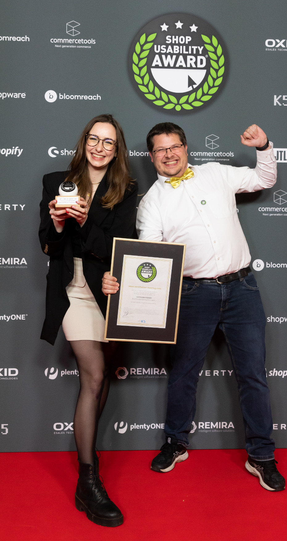 Zwei Personen feiern eine Auszeichnung auf einem roten Teppich. Eine Person hält eine Trophäe und eine Urkunde in den Händen. Im Hintergrund sind verschiedene Sponsorenlogos zu sehen.