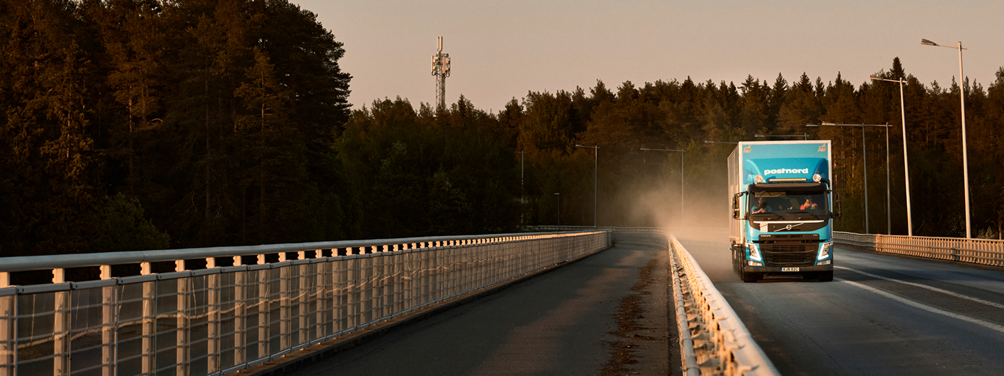 A blue PostNord delivery truck driving over a bridge surrounded by forest at golden hour, with a communication tower in the background.