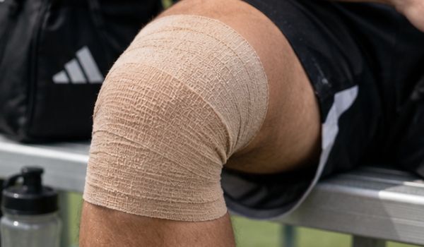 A knee wrapped in beige medical bandage, resting on a bench with a sports bag and water bottle nearby.