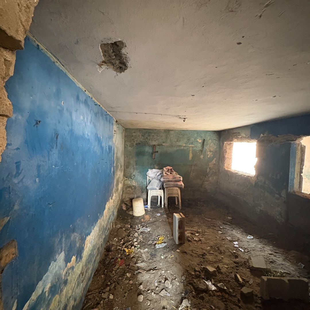 Dilapidated room with blue walls, a hole in the ceiling, debris on the floor, and two stacked chairs by a window with light streaming in.