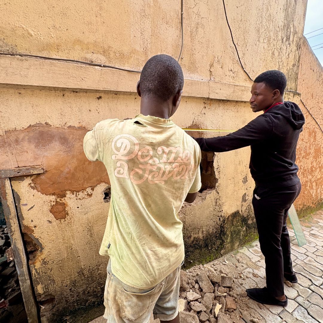 Two men working on a wall; one measures while the other stands nearby. They are outside on a cobblestone path.