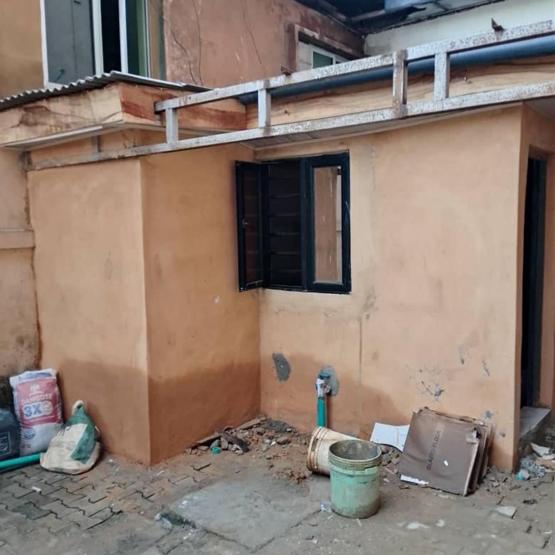 Small outdoor area with a tan plastered wall, window with black frame, scattered debris, and construction materials on the ground.