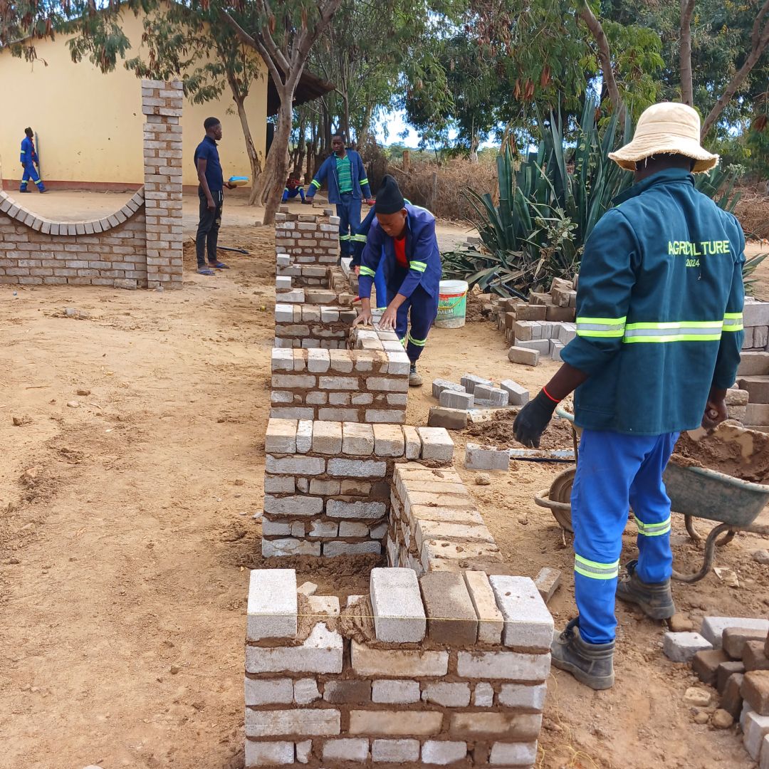 Workers in blue uniforms and hats are constructing a brick wall outdoors, surrounded by trees and a building.