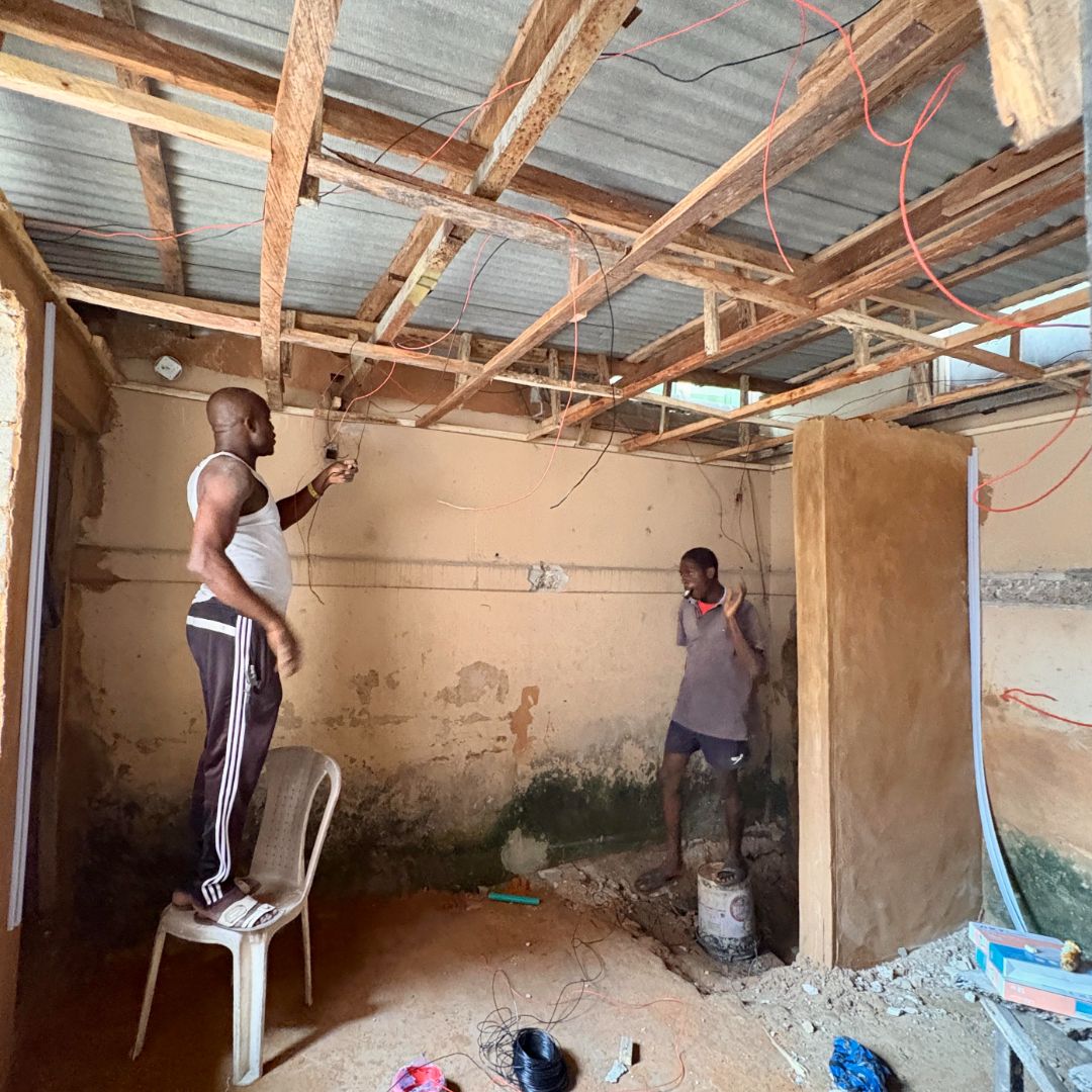 Two men work on electrical wiring in a room with exposed wooden beams. One stands on a chair, while the other stands on a bucket.