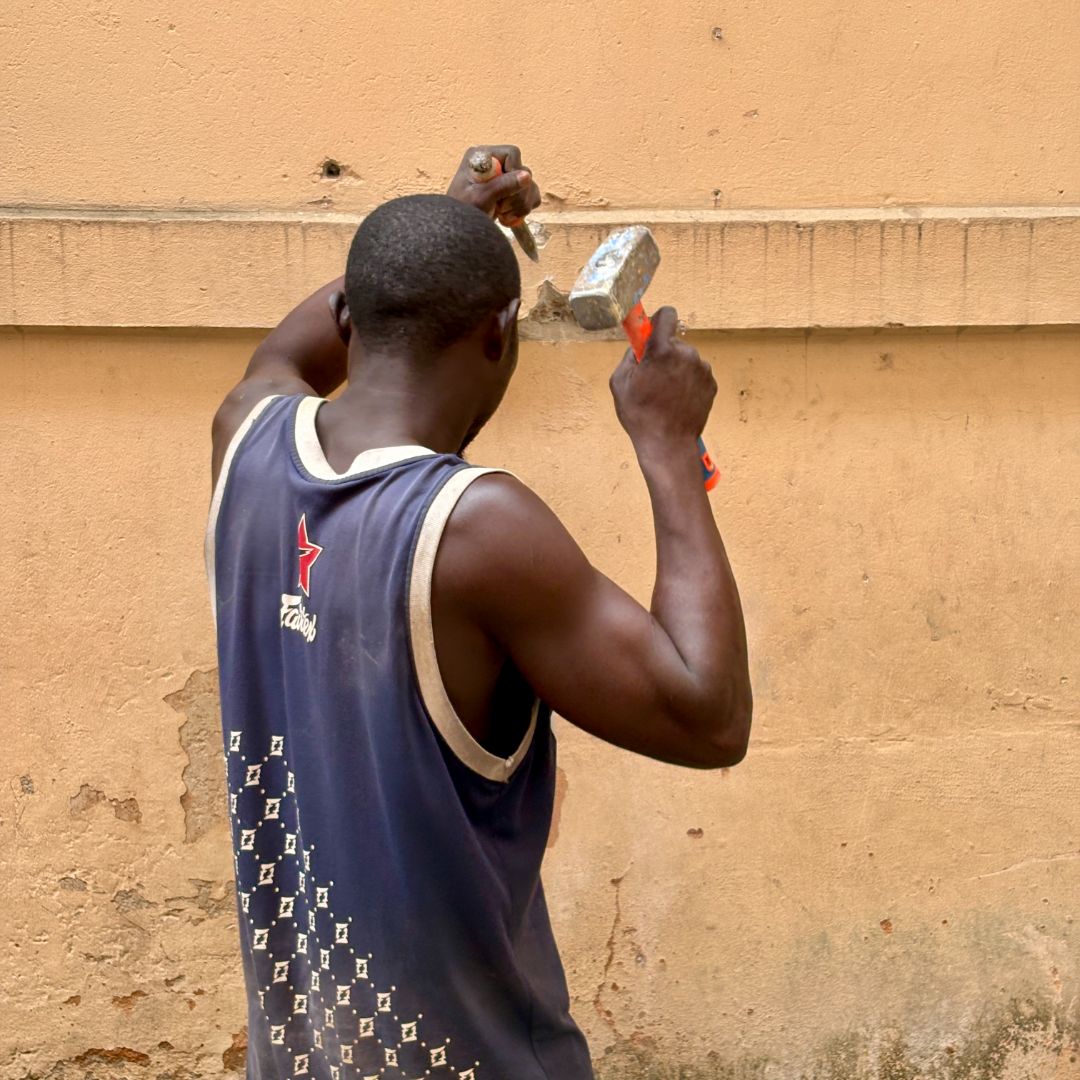 A man in a sleeveless shirt uses a hammer and chisel on a wall, focusing on his task.