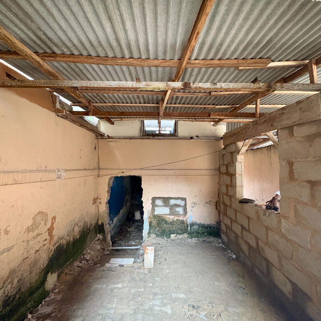 An empty, unfinished room with exposed corrugated metal roof, bare walls, a small window, and a doorway leading to another area.