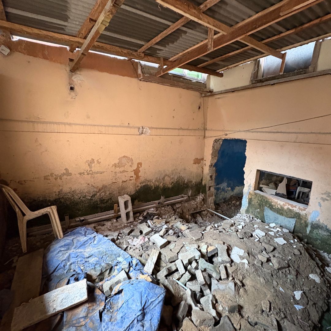 A dilapidated room with a partially collapsed ceiling, rubble on the floor, and a plastic chair against a moldy wall.