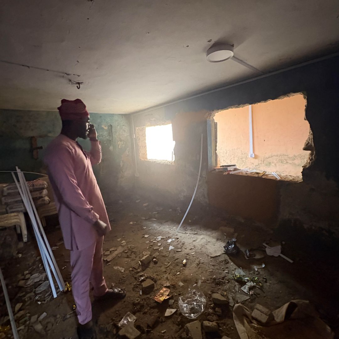A person in a pink outfit stands in a dim, cluttered room with a large hole in the wall, talking on a phone. Debris is scattered on the floor.
