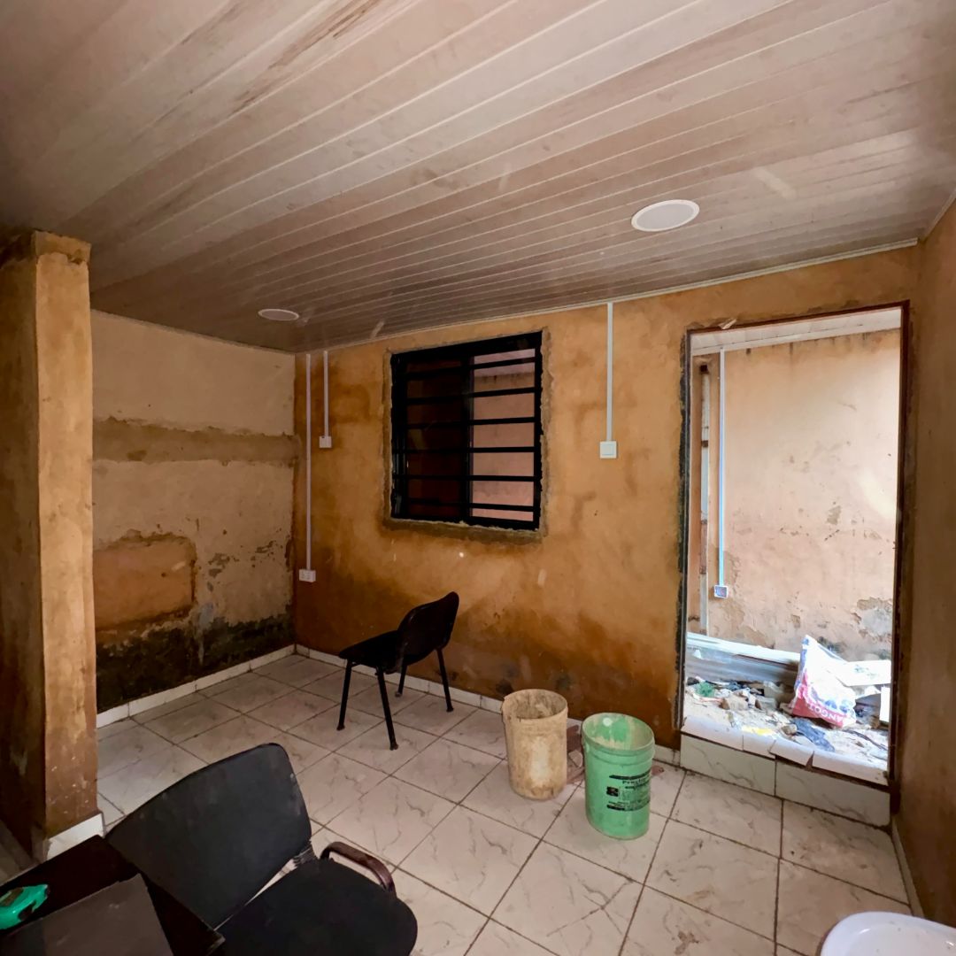 A small, unfinished room with exposed walls, a barred window, two chairs, and a bucket on tiled flooring. An open doorway leads outside.