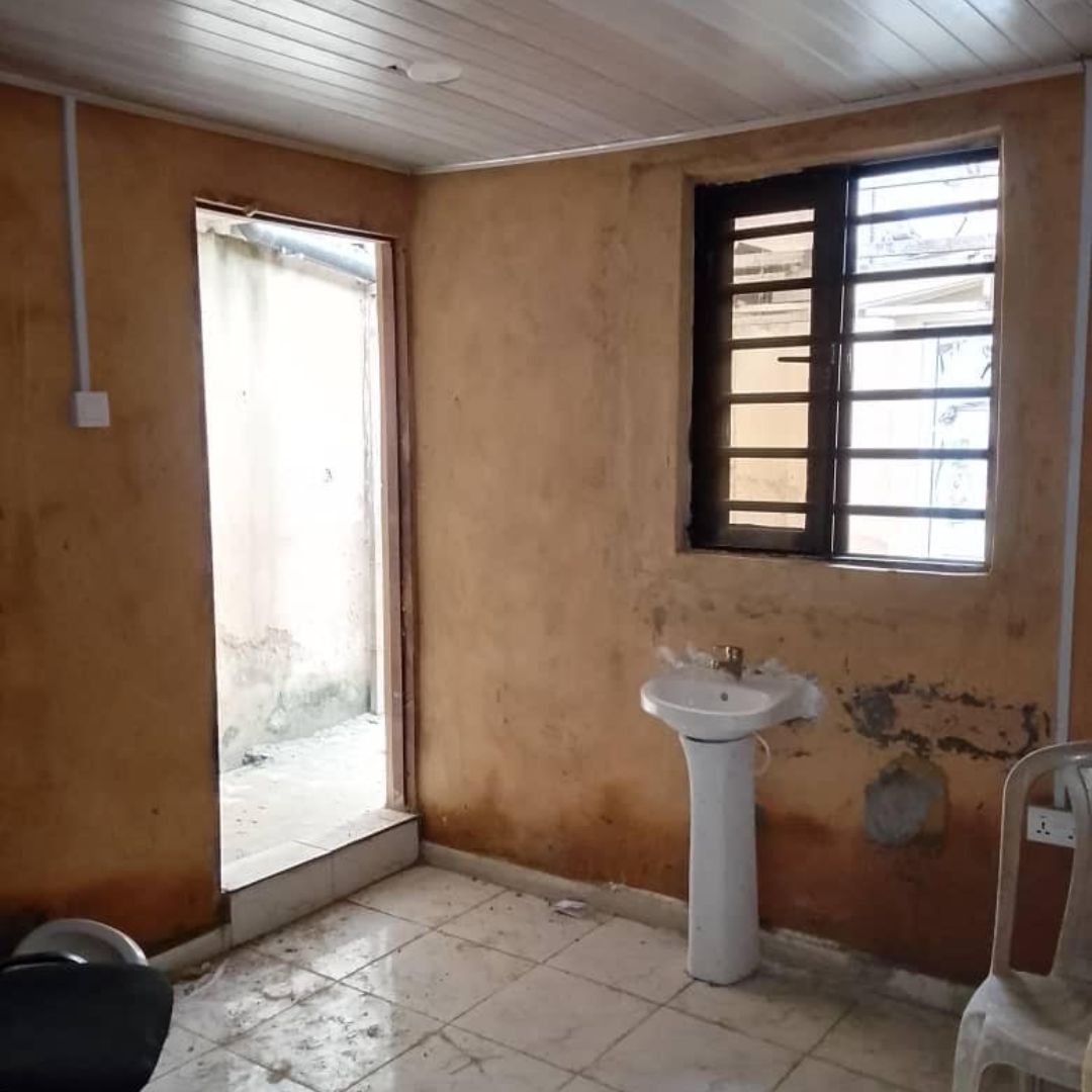 A small, unfinished room with a sink, open doorway, barred window, and tiled floor. Walls are unpainted with visible patches.