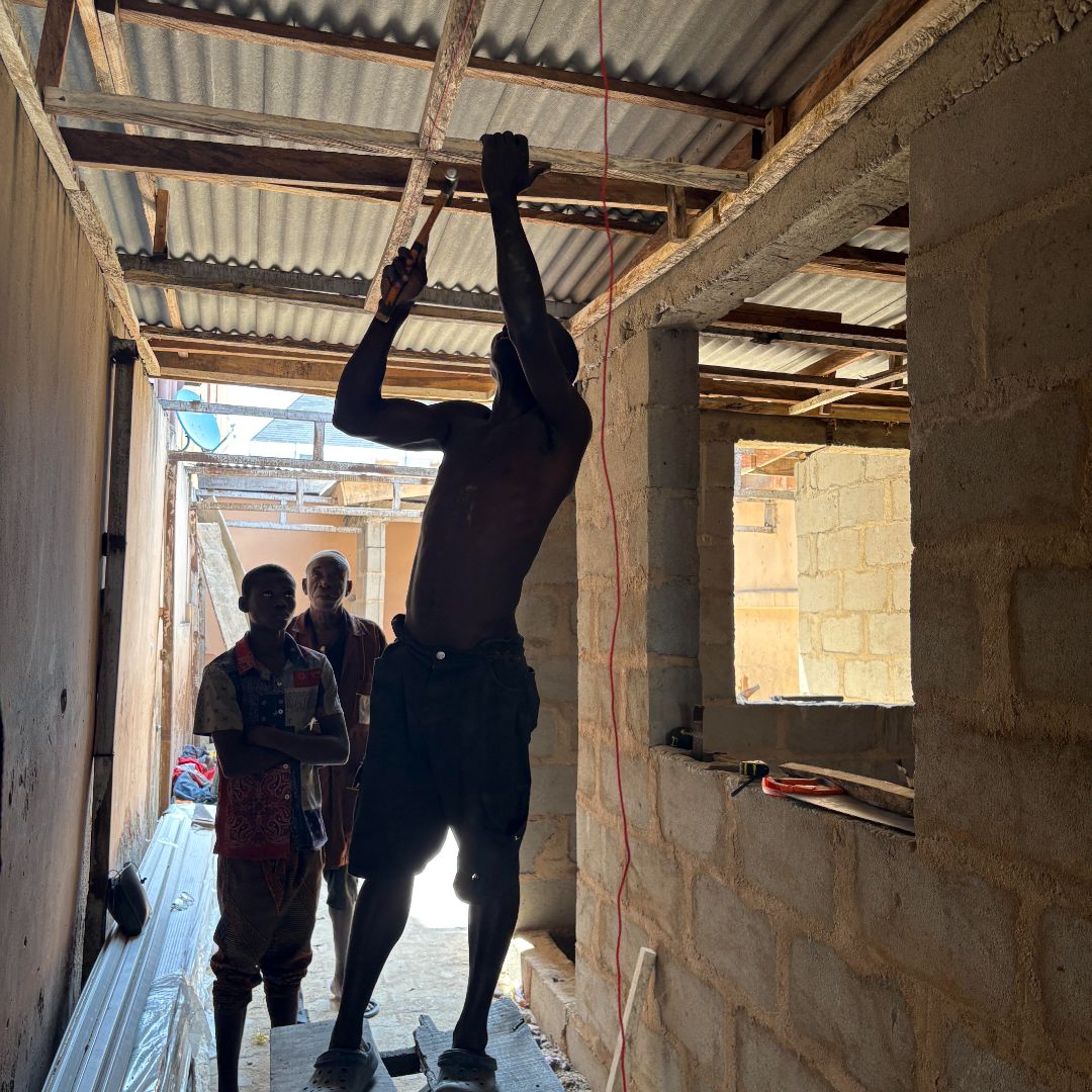 A person stands on a ladder installing a ceiling in a partially constructed building, with two people observing. Sunlight filters through the roof.