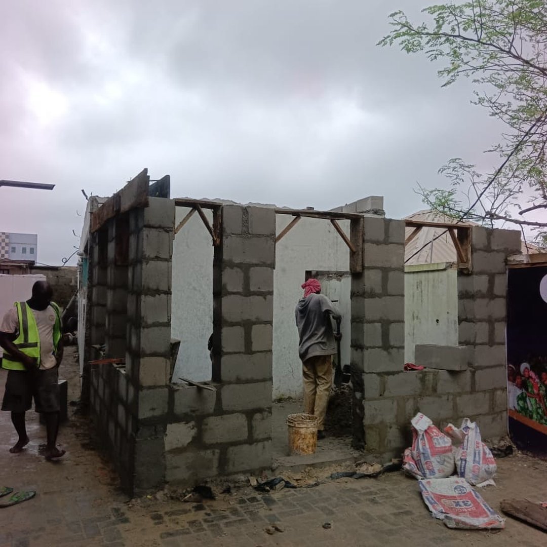 Two construction workers building a grey concrete block structure on a cobblestone street under an overcast sky.