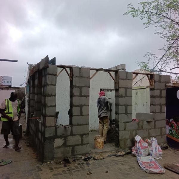 Two construction workers building a grey concrete block structure on a cobblestone street under an overcast sky.