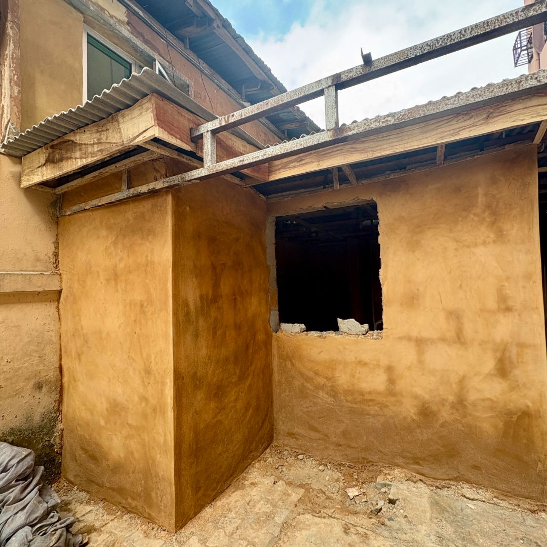 Partially constructed building with exposed wooden beams and an unfinished window opening, surrounded by a dirt floor and cloudy sky above.