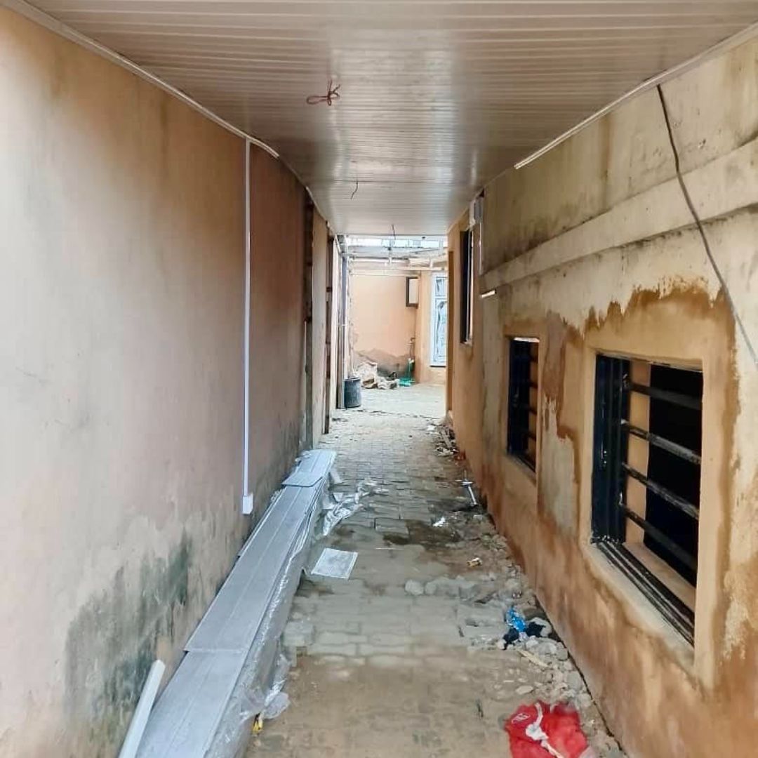 Narrow alleyway with beige walls, littered with debris and wires hanging. Sunlight at the far end.
