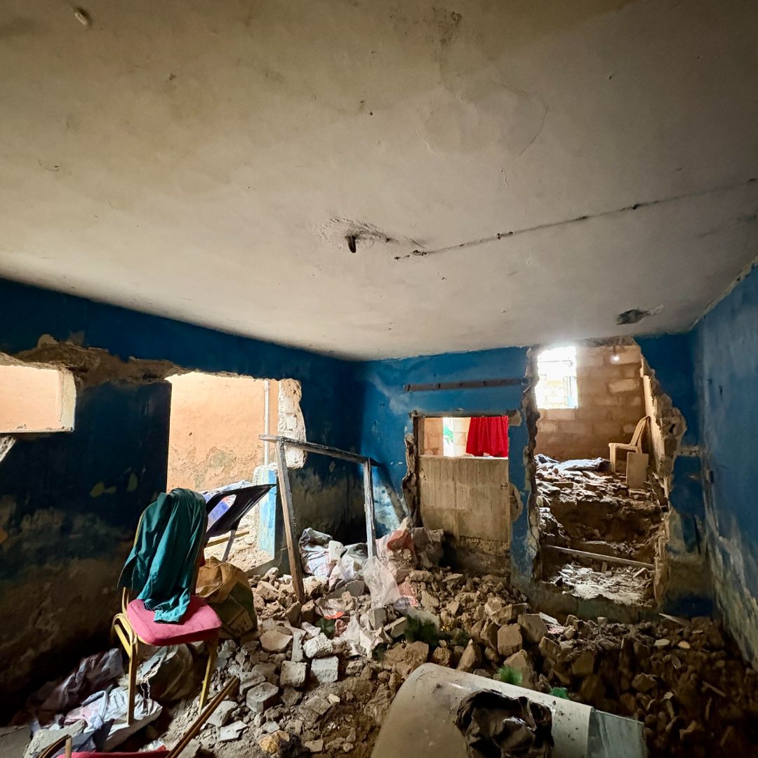 A dilapidated room with blue walls, debris on the floor, broken windows, and a partially collapsed wall leading to another room.