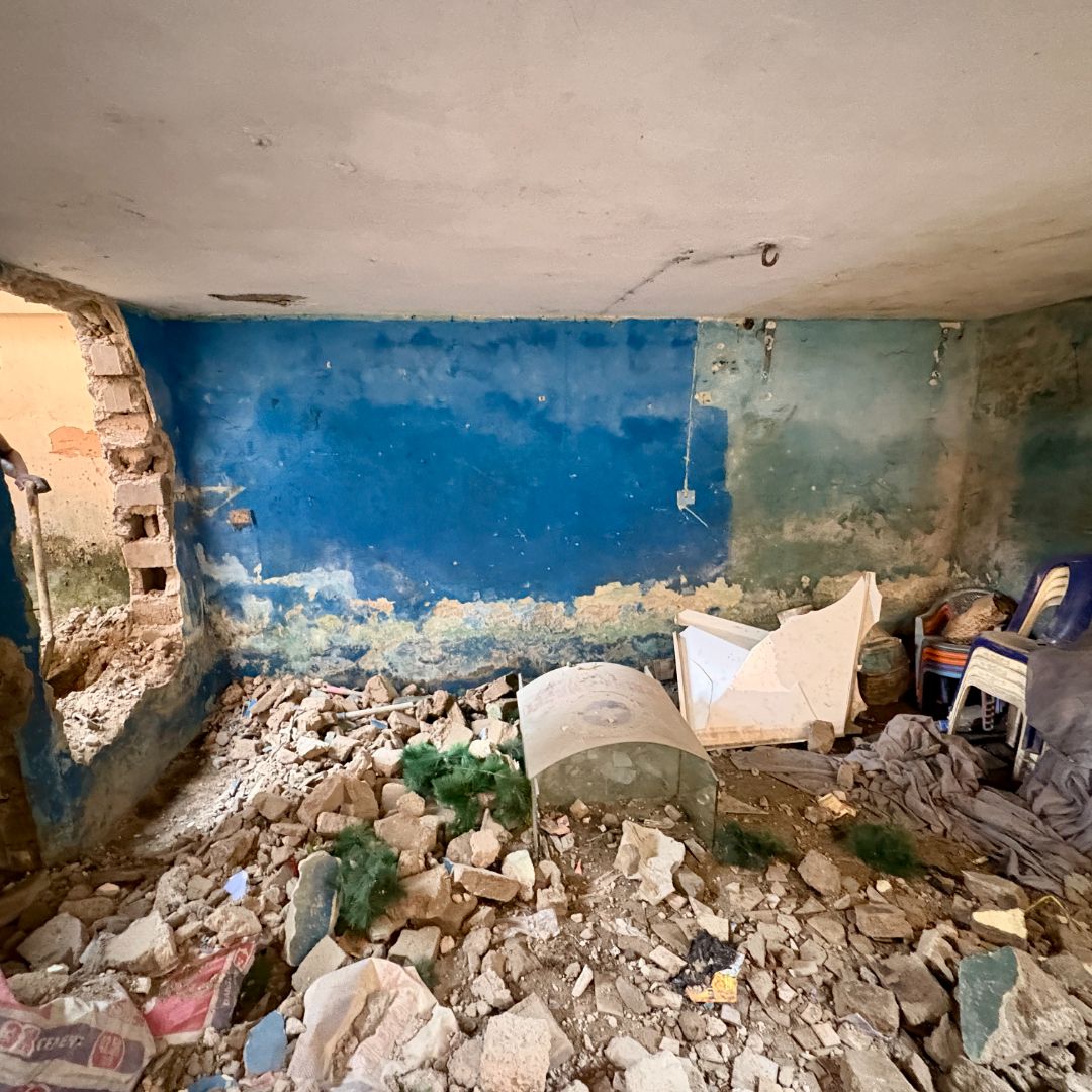 A dilapidated room with rubble on the floor, a damaged blue wall, broken furniture, and scattered debris.