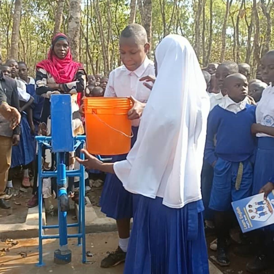 Students in uniforms operate a manual water pump, filling an orange bucket, surrounded by a group of onlookers in a wooded area.