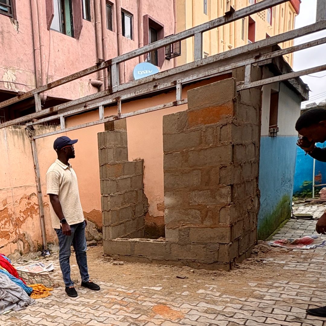 A man stands near a partially constructed brick structure in an alleyway, with surrounding buildings and scattered items on the ground.