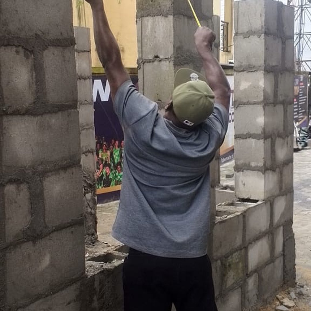 A man in a grey t-shirt and olive cap measuring a concrete block column at a construction site.