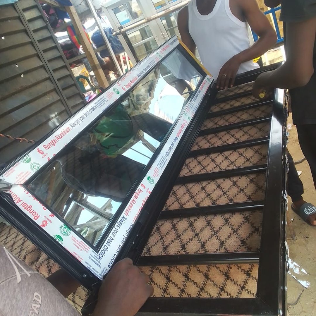 Workers handling a large black aluminium-framed glass window unit with protective Rongtai Aluminium branded tape in an open workshop setting.