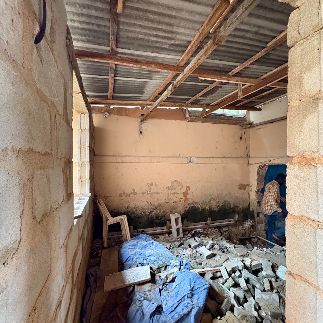 A partially constructed room with exposed brick walls, a metal roof, debris on the floor, and a person working in the corner.