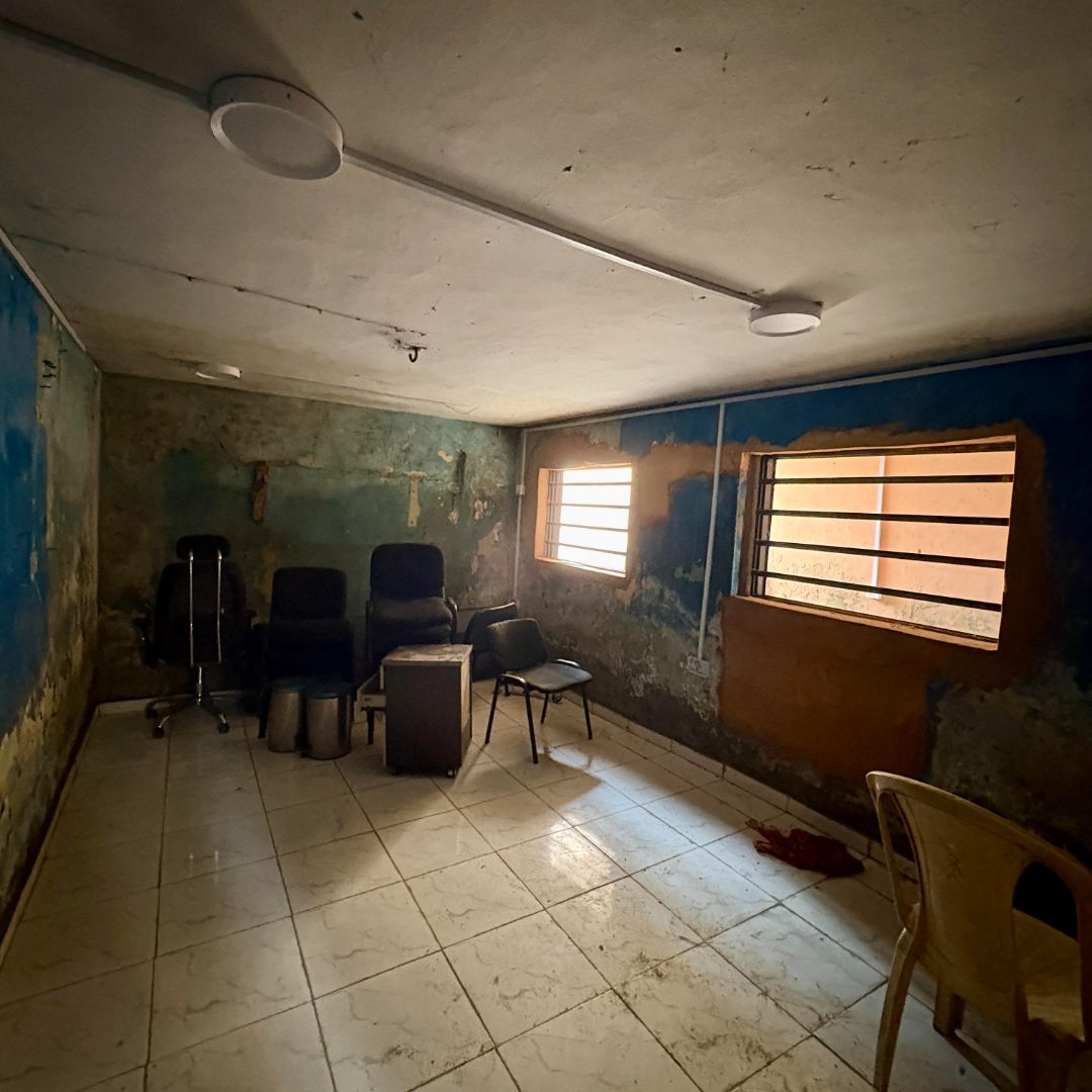 Dimly lit, worn-out room with peeling paint, chairs, a small table, and barred windows. The floor is tiled, and the walls are blue and green.