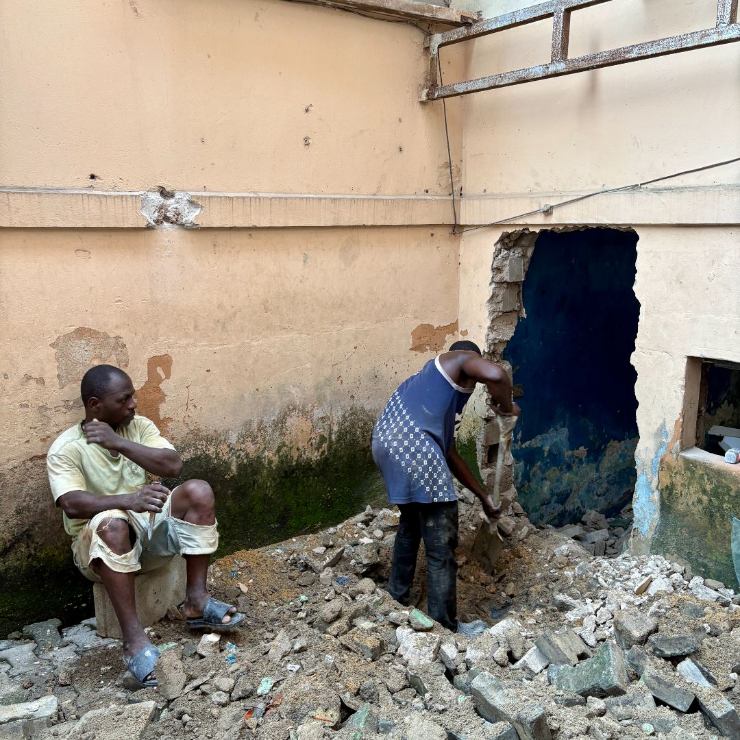 Two people work in a damaged building. One sits on rubble, while the other clears debris from a large hole in the wall.