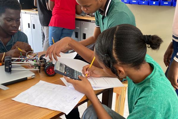Students in green and red shirts working on a robotic vehicle project with a calculator and handwritten notes on a wooden table.