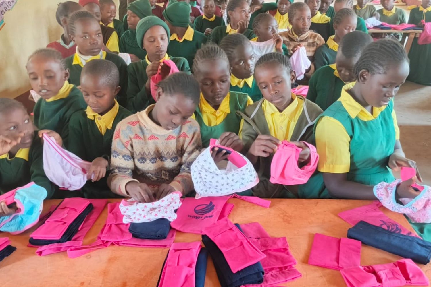 Children in school uniforms sit at a table, examining colorful reusable sanitary pads and underwear, with more supplies laid out in front of them.