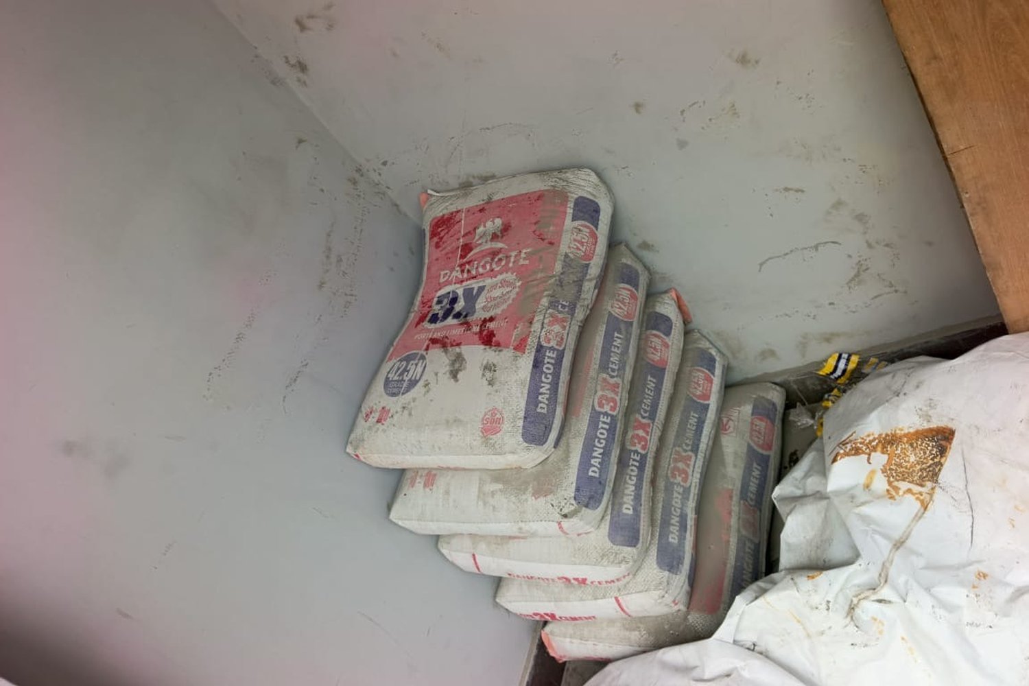 Bags of Dangote 3X cement stacked in the corner of a grey-walled storage space alongside other white sacks.